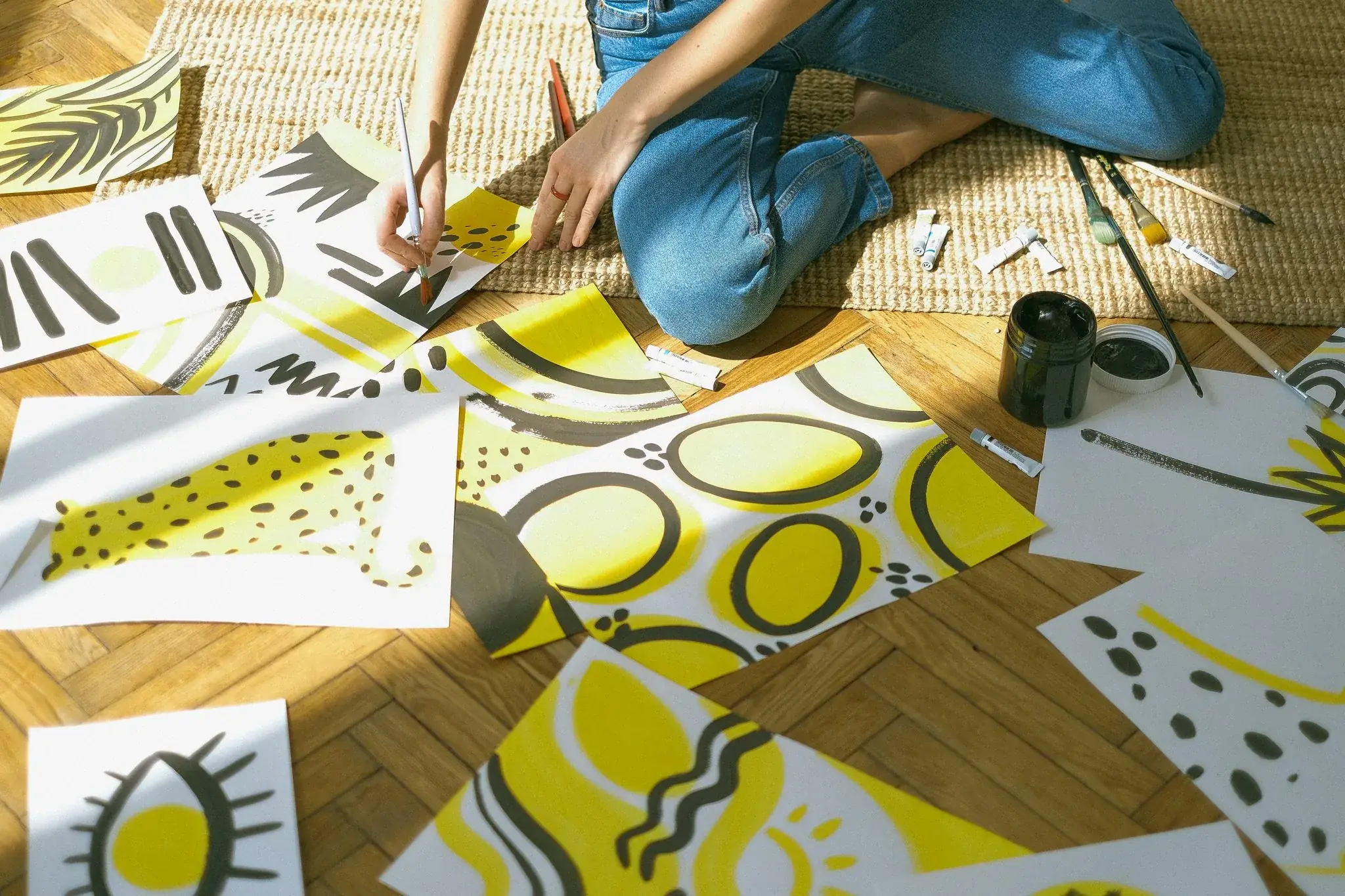 Artist seated on the floor painting bold yellow and black abstract patterns across scattered sheets of paper