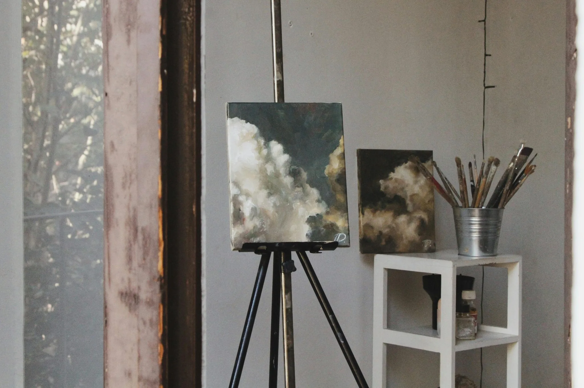 Oil paintings of dramatic clouds on an easel and shelf in a sunlit art studio