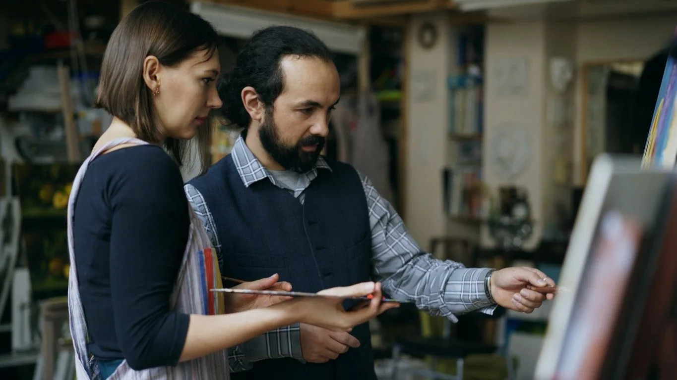 Art mentor pointing at a canvas while a student holding a palette looks on in a painting studio
