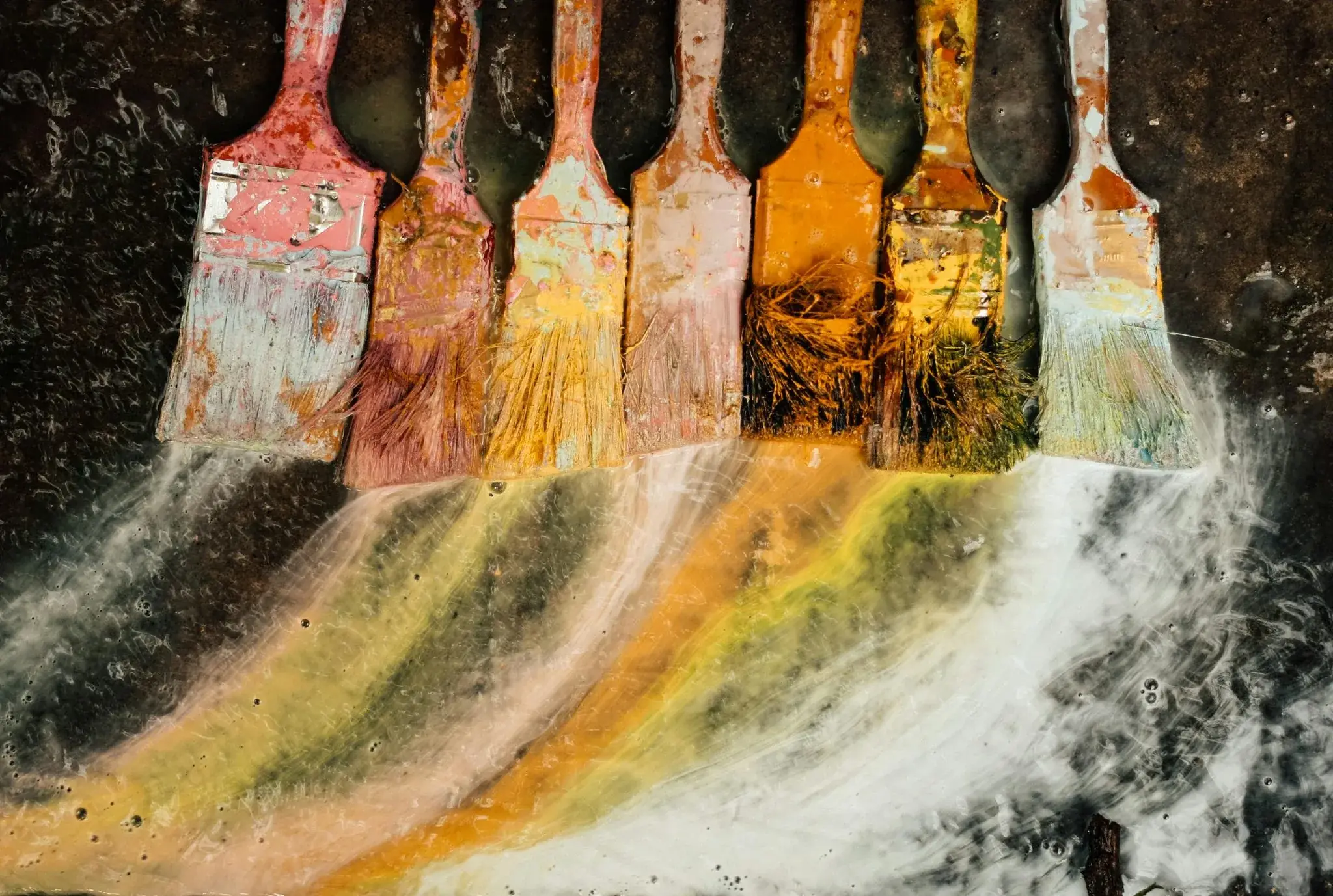 Row of paint-crusted brushes smearing streaks of red, yellow and green across a dark surface
