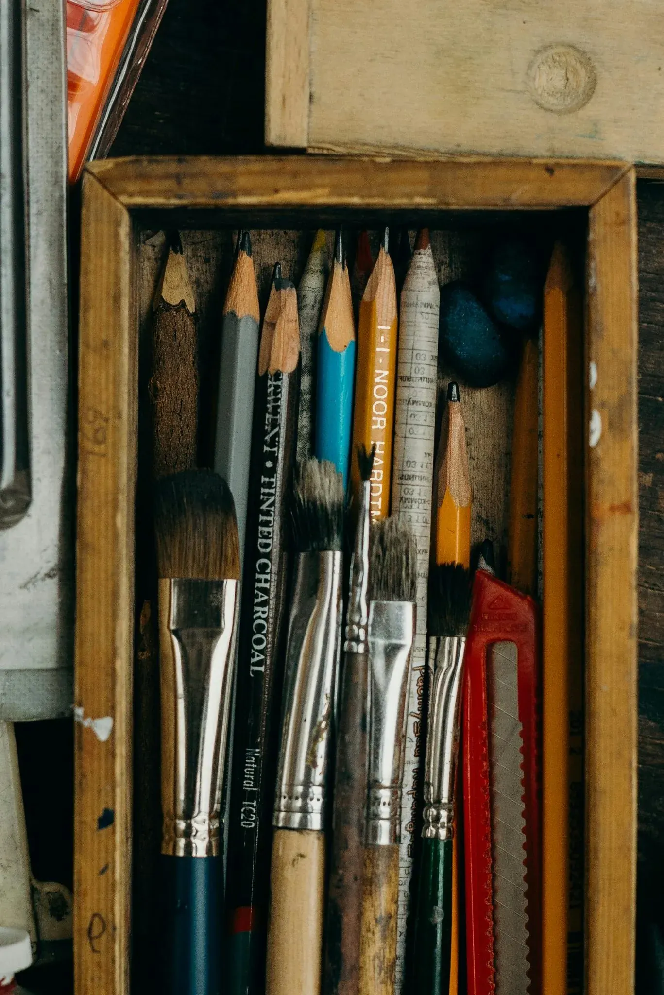 Well-used paintbrushes, charcoal and coloured pencils stored upright in a worn wooden box