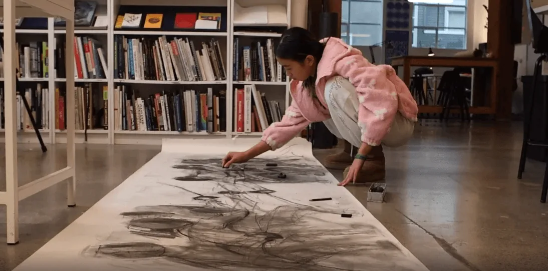 Student reaching across a long studio table to work on a large charcoal drawing beside bookshelves