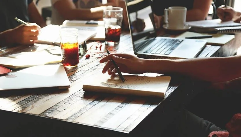 Student taking handwritten notes in a notebook beside laptops during a study session
