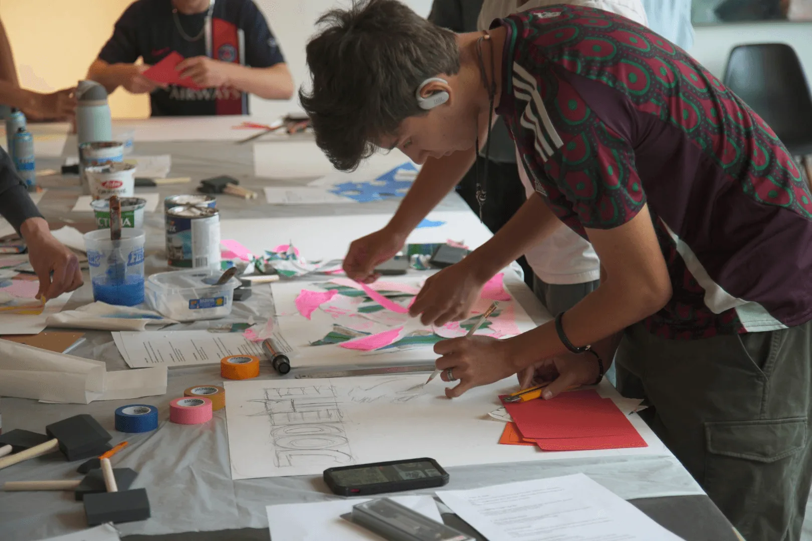 Teen students cutting paper and lettering posters at a paint-covered workshop table