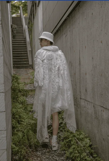 Model wearing a translucent white ruffled cape and bucket hat on an overgrown concrete stairway