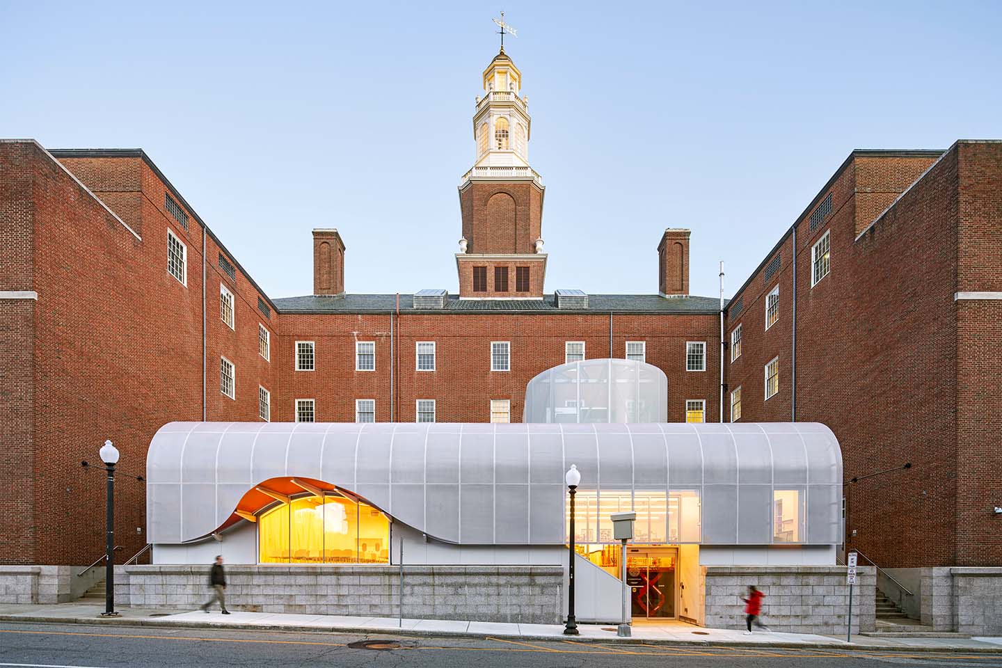 Historic brick university building with clock tower behind a curved translucent modern pavilion at street level