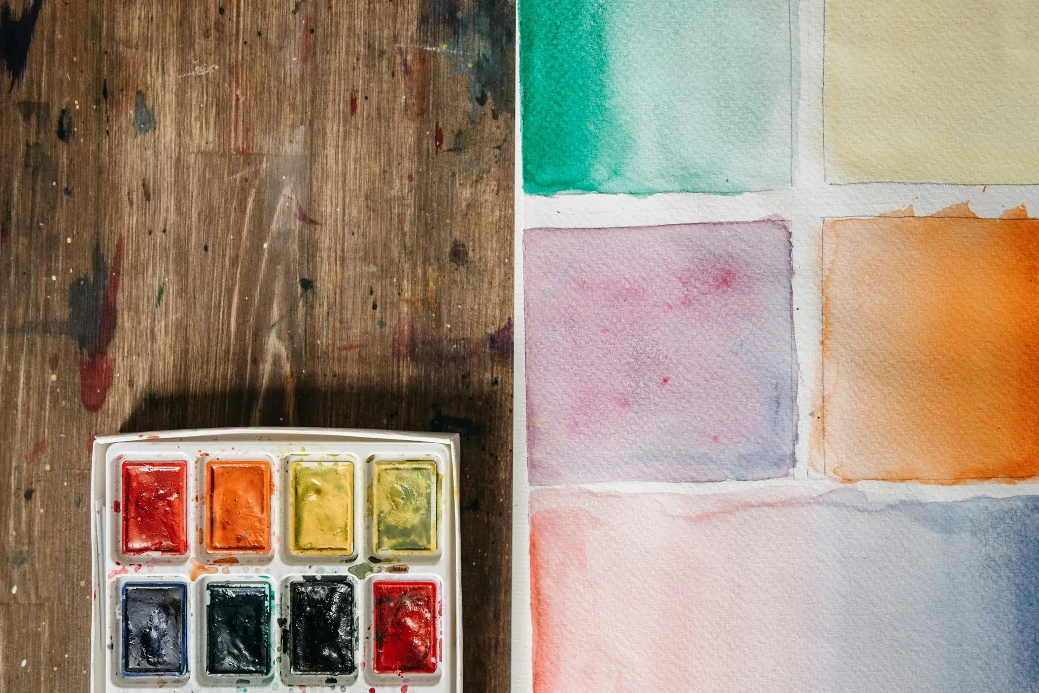 Watercolour palette on a paint-splattered wooden desk beside a sheet of colourful test swatches